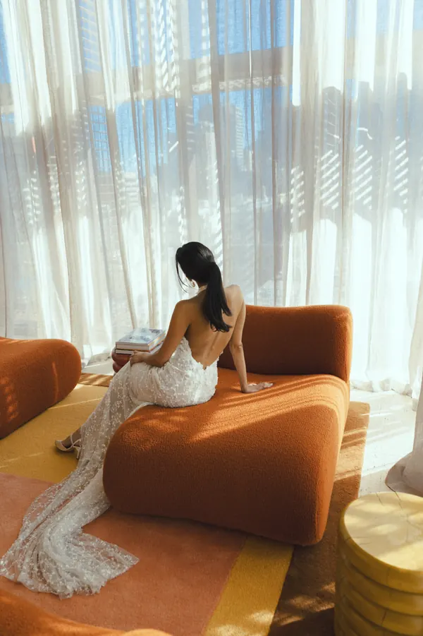 Seated by a sunlit window in a lace gown, dark hair in a sleek low ponytail