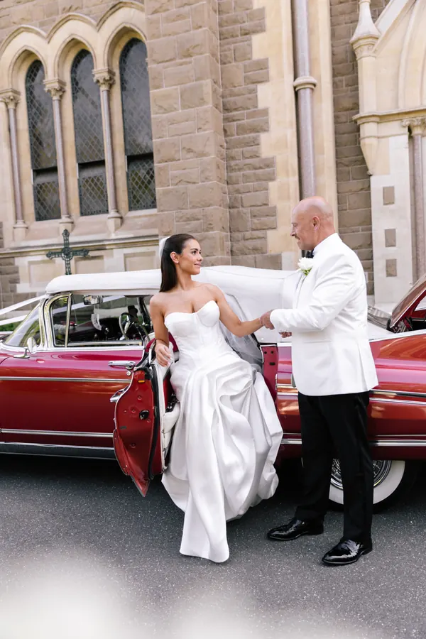 Stepping out of a vintage car outside a bluestone church, hair worn long and straight